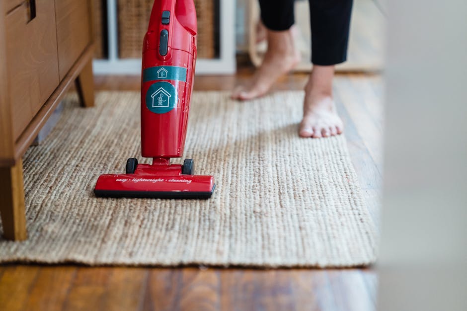 Close-up of a person using a yellow and black portable vacuum cleaner to perform surface cleaning on a patterned carpet in a domestic setting. The individual is wearing blue jeans and a beige top, hol